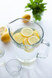 A glass pitcher filled with fresh lemon water, surrounded by lemons and mint, on a bright table.