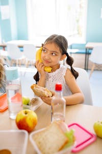 A young girl playfully holds a banana while enjoying a sandwich at a classroom table.